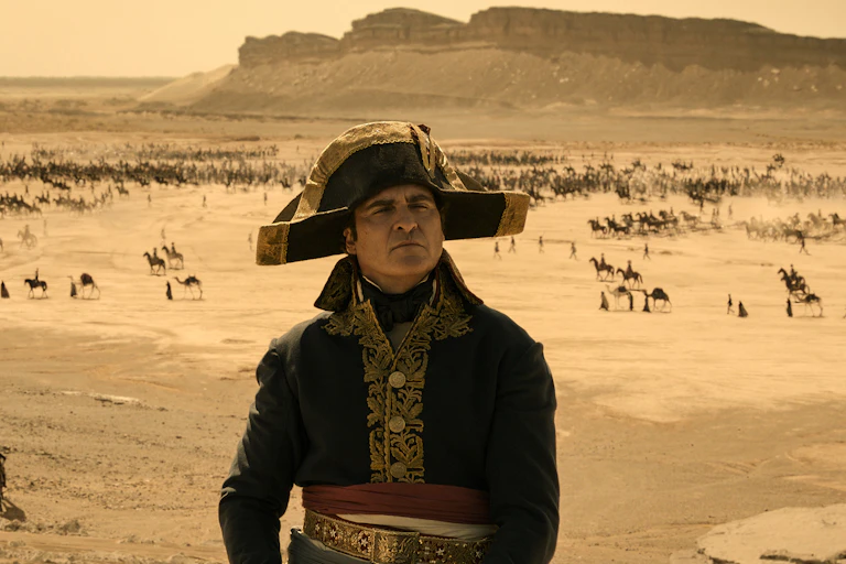 o ator Joaquin Phoenix caracterizado como Napoleão num deserto com camelos passando ao fundo.