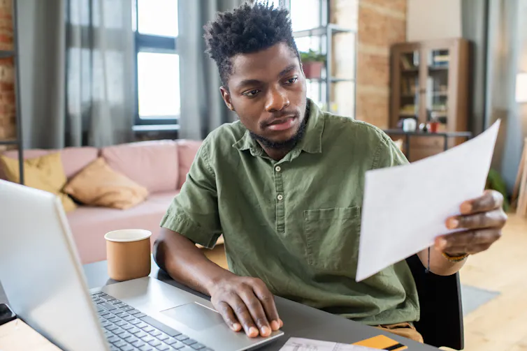 A young man sitting at a laptop computer, looking in surprise at a piece of paper