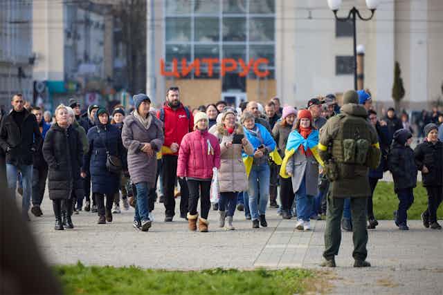 People walk wearing hats and coats walk toward Ukraine's Volodymyr Zelensky, back to camera.