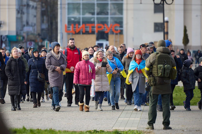People walk wearing hats and coats walk toward Ukraine's Volodymyr Zelensky, back to camera.