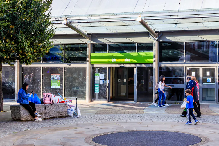 An outdoor plaza in front of a building housing a Jobcentre office, with people and a tree.