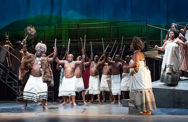 A stage full of bare chested men in traditional skirts holding sticks aloft while women in Xhosa traditional attire sing and dance to one side.