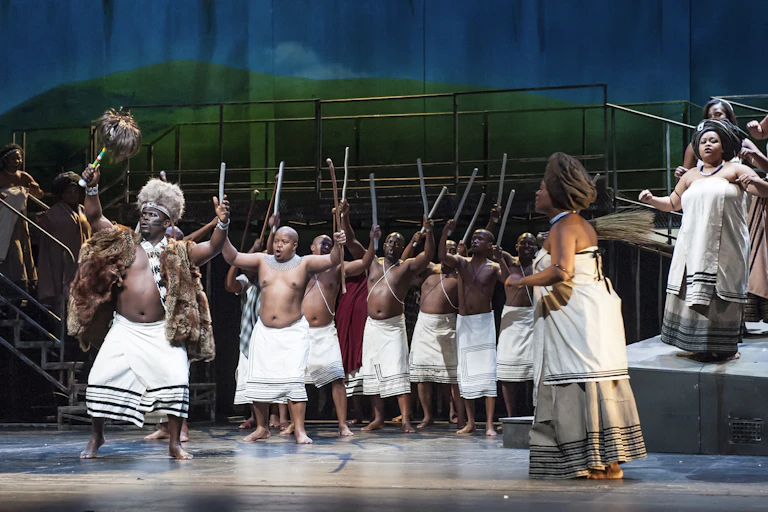 A stage full of bare chested men in traditional skirts holding sticks aloft while women in Xhosa traditional attire sing and dance to one side.