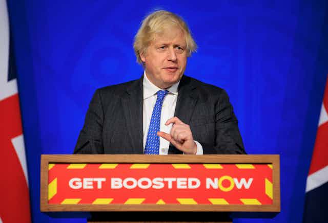 Boris Johnson speaking at a podium that reads 'get boosted now' during a coronavirus update