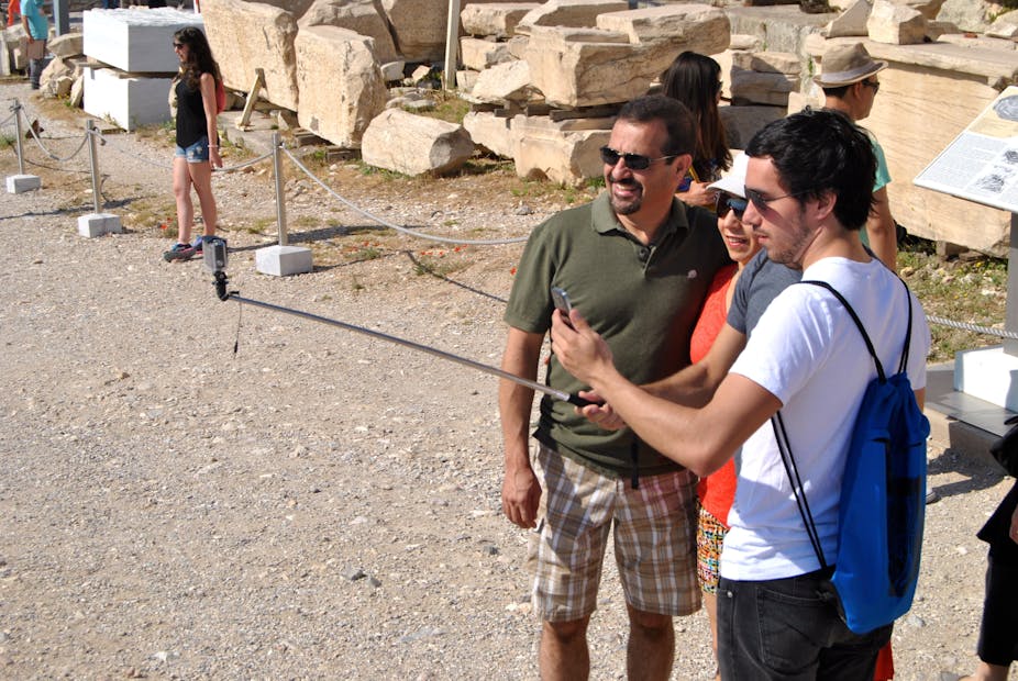 Three tourists look at a phone on a selfie stick in front of stones from the Acropolis in Greece.