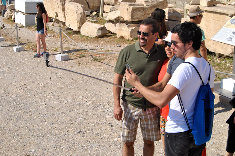 Three tourists look at a phone on a selfie stick in front of stones from the Acropolis in Greece.