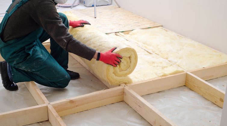 A worker in overalls unrolls a yellow insulating foam in a wooden lattice.
