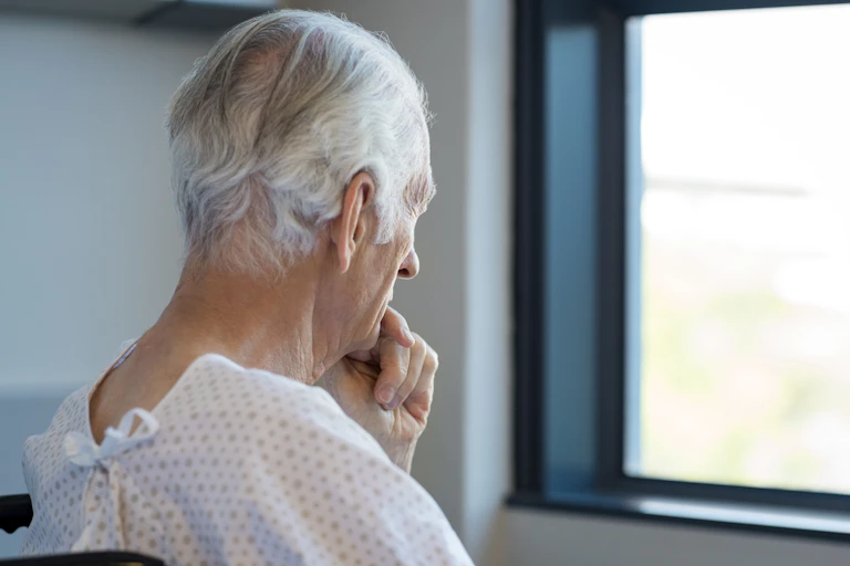 Photo d’un homme âgé en tenue d’hôpital en train de regarder pensivement par la fenêtre en se tenant le menton.