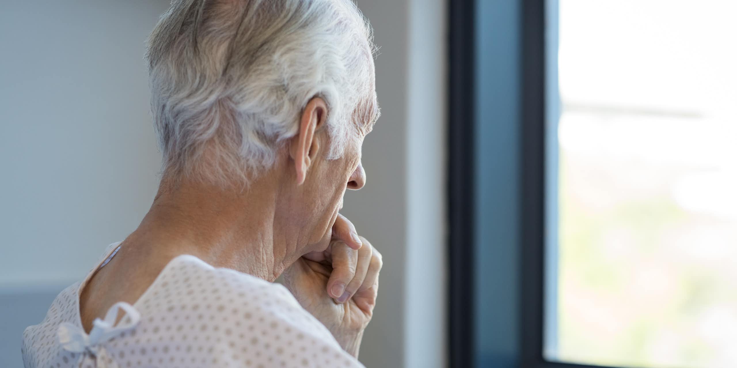 Photo d’un homme âgé en tenue d’hôpital en train de regarder pensivement par la fenêtre en se tenant le menton.