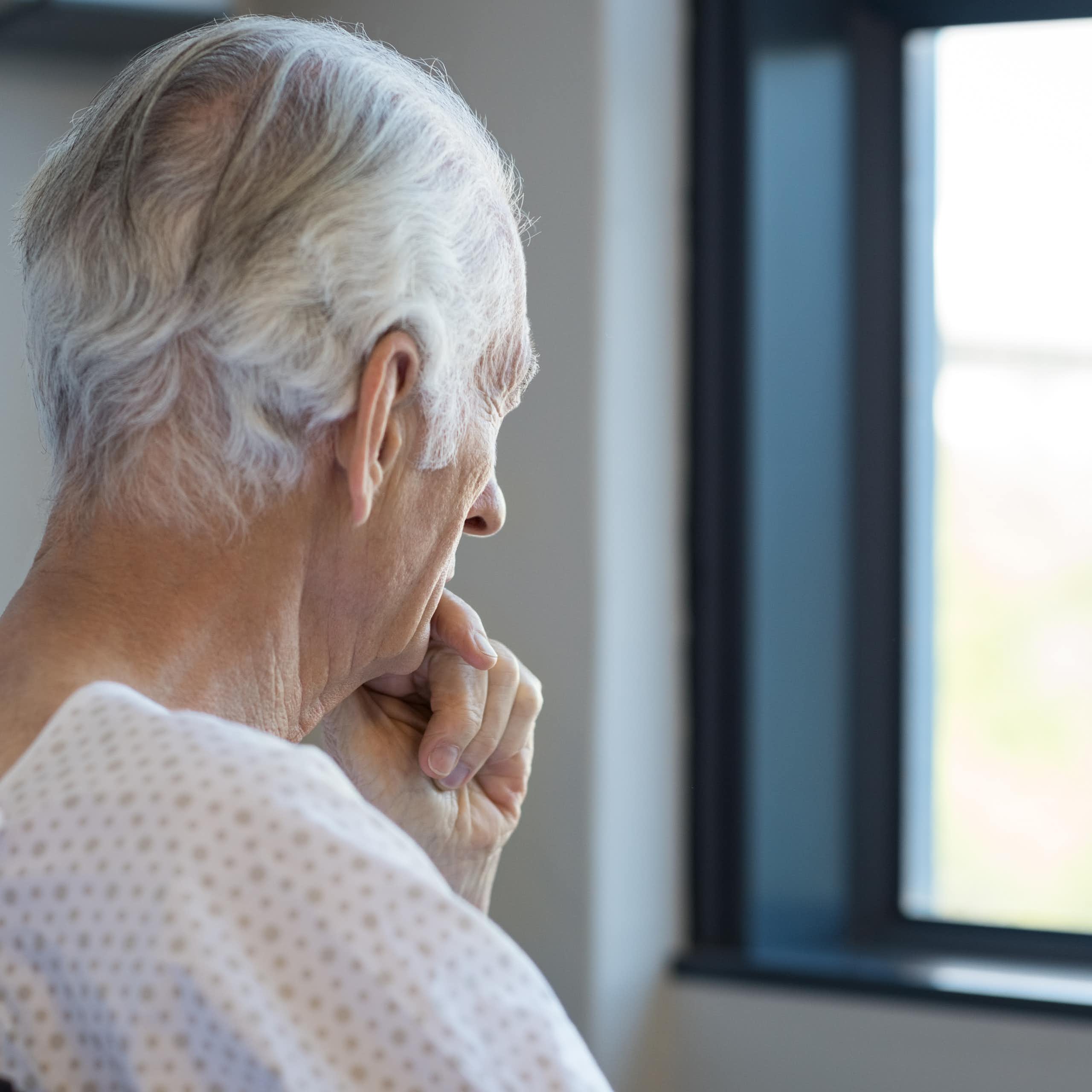 Photo d’un homme âgé en tenue d’hôpital en train de regarder pensivement par la fenêtre en se tenant le menton.
