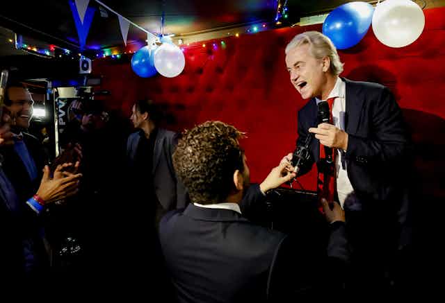 Geert Wilders on stage with a microphone with a crowd smiling and taking pictures in front of him. 