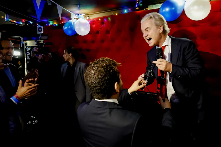 Geert Wilders on stage with a microphone with a crowd smiling and taking pictures in front of him.