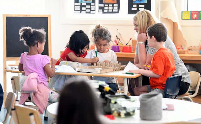 An early childhood educator sits at a table with four child children doin a craft activity.