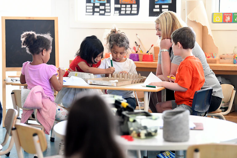An early childhood educator sits at a table with four child children doin a craft activity.