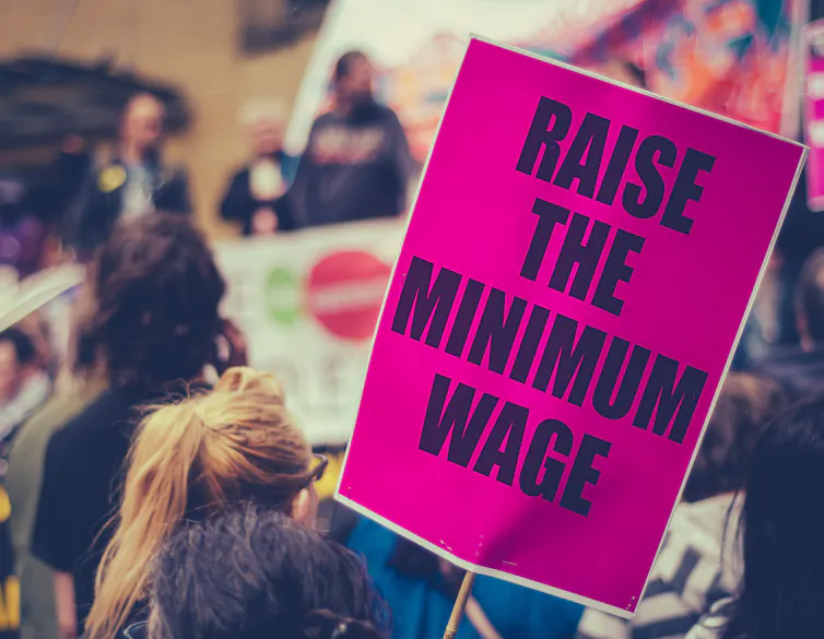 Person at a protest with a pink sign that says raise the minimum wage.