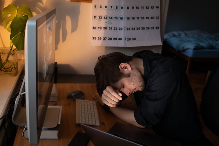 A man at a home working desk rests his head on the desk in front of his computer