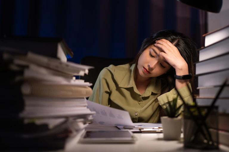 A young woman sitting at a desk in the dark, piled high with books and papers