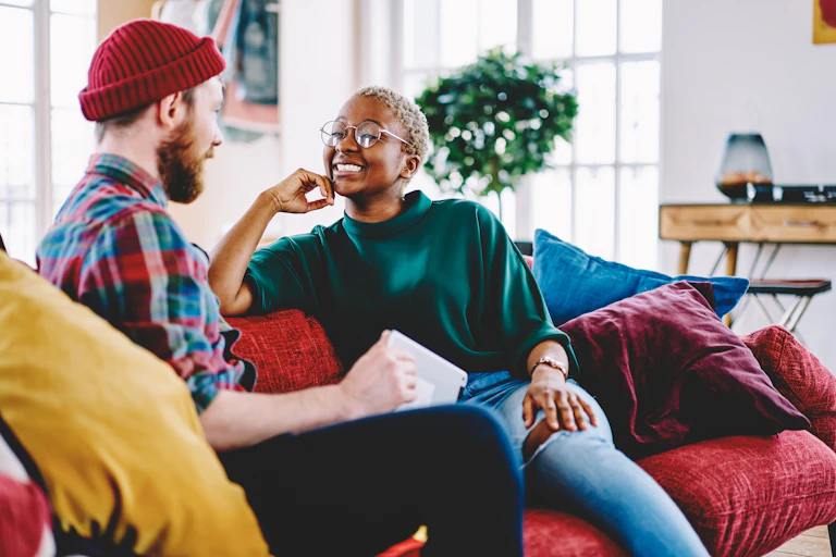 A young couple sits on a couch in a colourful, modern apartment, smiling at each other, engaged in conversation