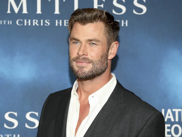 Actor Chris Hemsworth wearing a dark suit jacket and white shirt with open collar in front of a blue backdrop that reads 'Limitless with Chris Hemsworth'