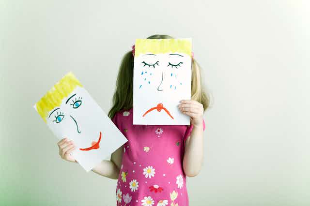Uma garota loira segura um desenho de um rosto triste na frente de seu rosto e um desenho feliz em sua mão