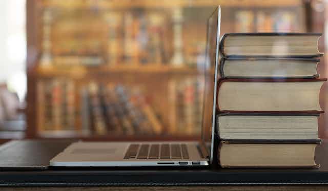 A stack of five books sits next to an open laptop on a table in a library.