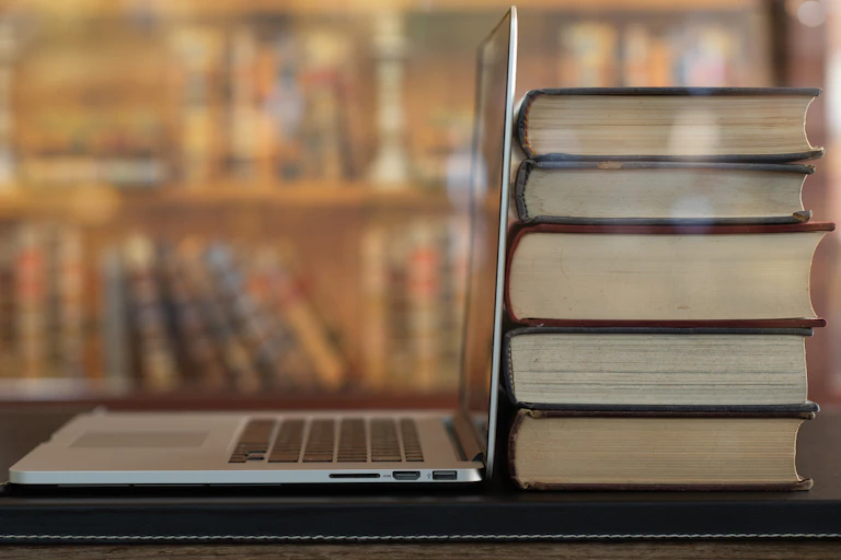 A stack of five books sits next to an open laptop on a table in a library.