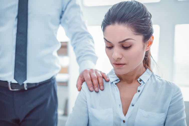 Seated woman looking uncomfortable as a man in a suit rests his hand on her shoulder