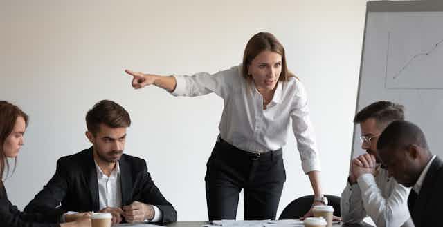 Woman gesticulating in a threatening manner to a man a sitting at a table with a group of colleagues