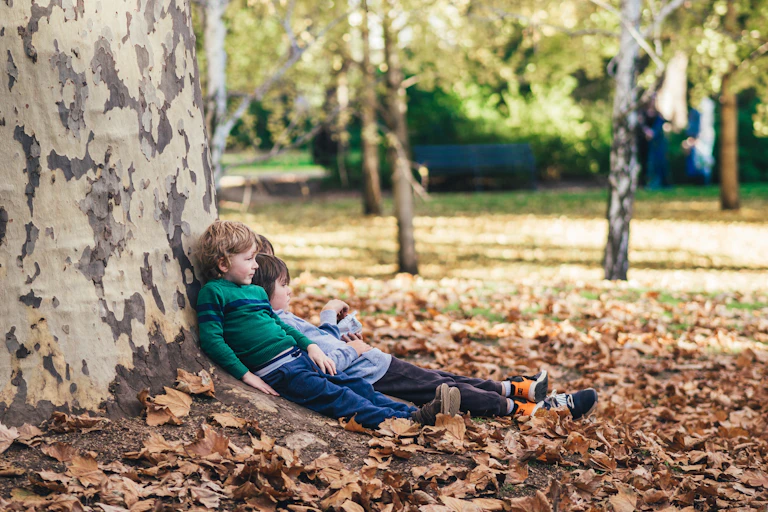 Small children sit propped up against a large tree in a park.