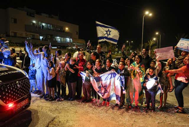 A crowd of around two dozen people cheer, some holding Israeli flags, as a car drives past at night.