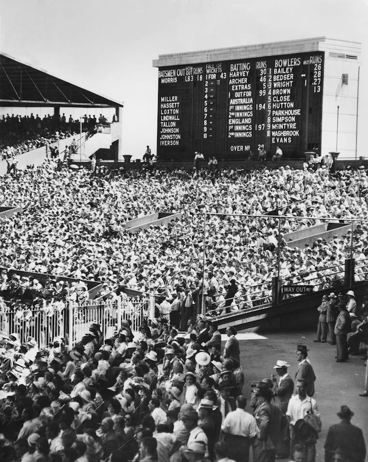 A black and white scene of dense crowds gathering in front of a cricket scoreboard in 1950