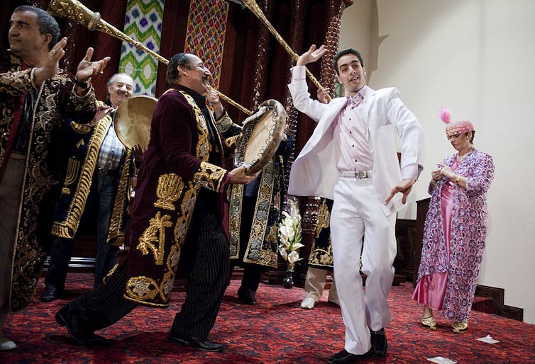 A young man in a white suit dances on a red carpet as older men in ornate robes play instruments around him.