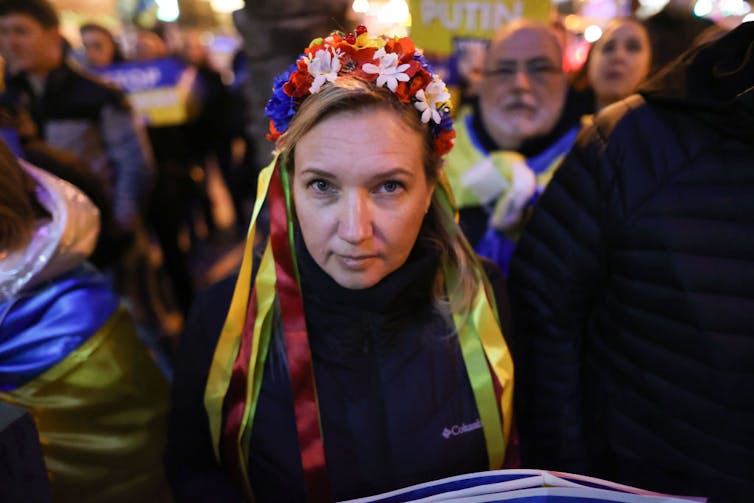 A woman with yellow ribbons and red and white flowers in her hair looks solemnly at the camera amid a protest.