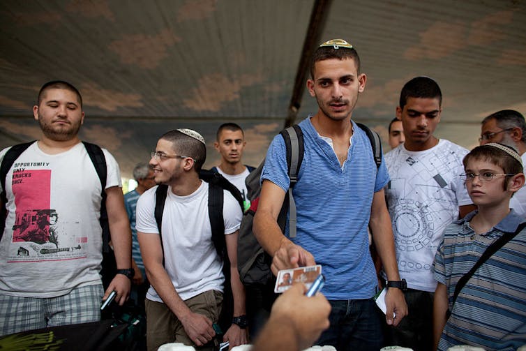 A handful of young men in t-shirts crowd around a table, unseen, to receive laminated cards.