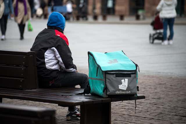 Man with blue hood and black coat sitting on a bench beside a blue and grey Deliveroo delivery bag.