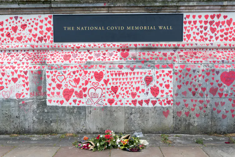 The UK's national covid memorial wall in London, a concrete wall covered in illustrated red hearts, with a few bouquets of flowers at the base.