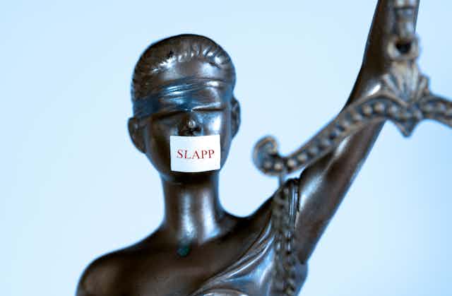 A blindfolded statue representing justice but with a sticker over he mouth bearing the word 'SLAPP'.