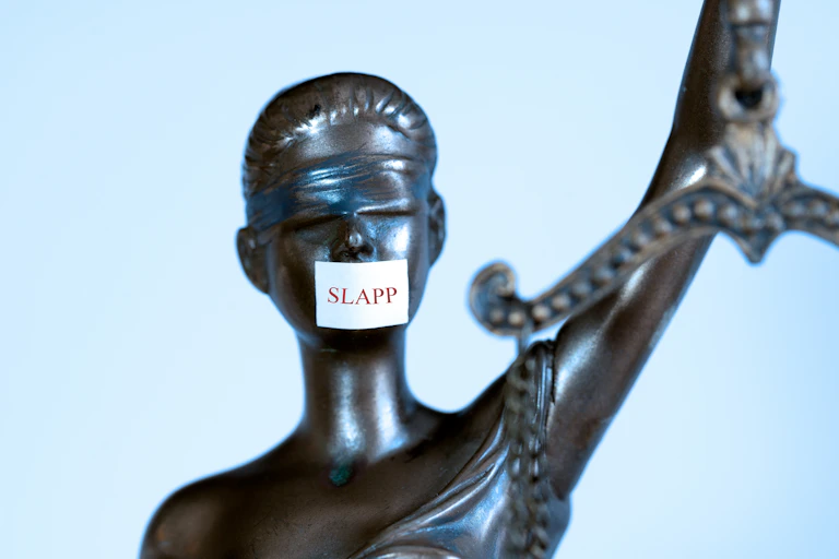 A blindfolded statue representing justice but with a sticker over he mouth bearing the word 'SLAPP'.