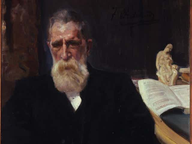 Un hombre de barba blanca se sienta al lado de una mesa con un libro abierto.