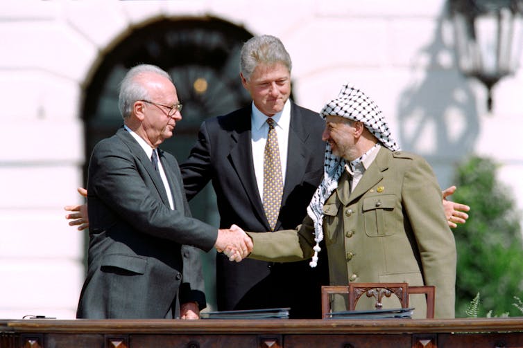Two men shake hands while a third stands between them, smiling.