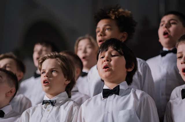 Children wearing white shirts and black bowties sing in a choir.