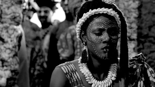 A woman stands in the foreground, watched by a man in the background. She wears shells in her hair and around her neck and has her face painted in white dots.