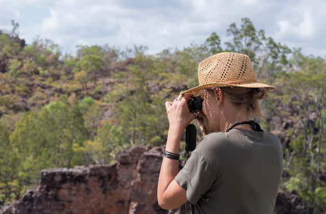 A woman in a sun hat looks at the view through a pair of binoculars near Jabiru in Kakadu National Park in Australia