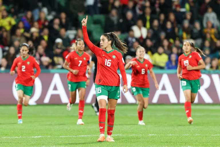 A woman in red and green sports clothing raises her hand, pointing a finger in victory. Behind her four other players run towards her and behind them a packed stand of people.