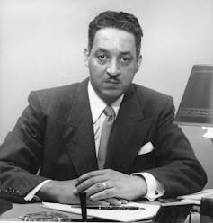 A Black man dressed in a business suit is sitting behind a desk with his hands crossed.