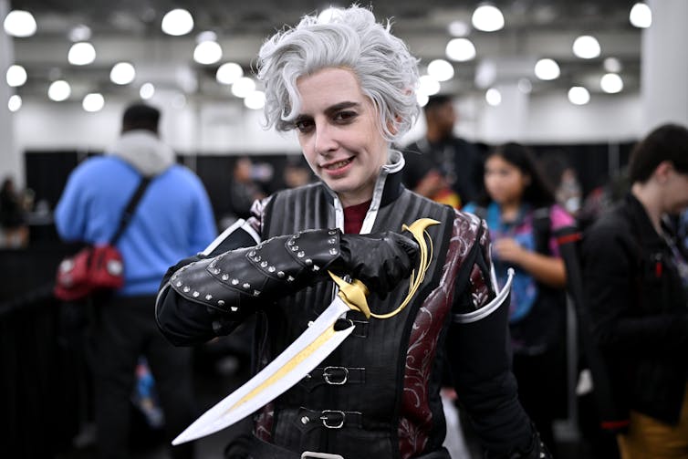 Young man with hair dyed white and gray grins as he poses with a dagger.