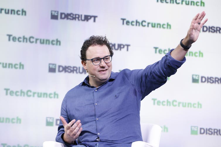 Man in blue button-down shirt gesticulates with one arm outstretched against a backdrop with the words TechCrunch and DISRUPT