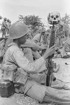 Un joven soldado vestido de uniforme sostiene un cráneo humano encima de su rifle.