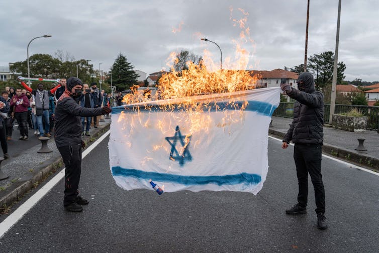 Two hooded men burning a white and blue Israeli flag.