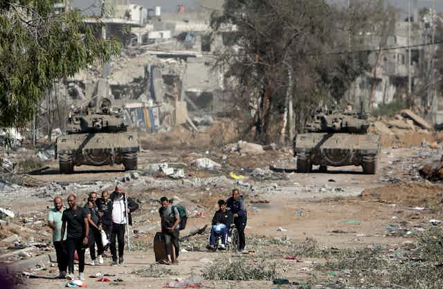 People walk along a road with tanks behind them pointing in their direction. Destroyed buildings seen in the backgroung